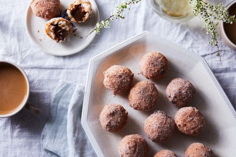 Ten brown, cinnamon-covered balls sit on an octagonal white plate. Three more are on a small, round white plate. There's a bite out of one, and you can see creamy brown Nutella inside. A cup of coffee with cream peeks in from the left.
